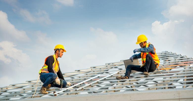 Two roofers in yellow helmets and safety vests install metal roofing panels on a house.