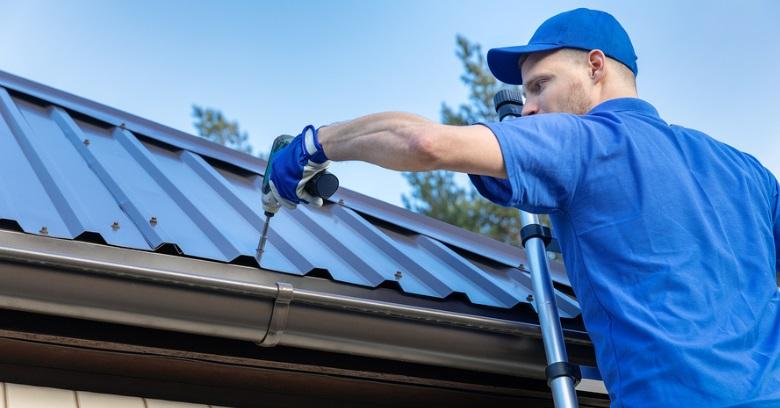 Roofing worker in blue uniform installing metal roofing panels on a house.